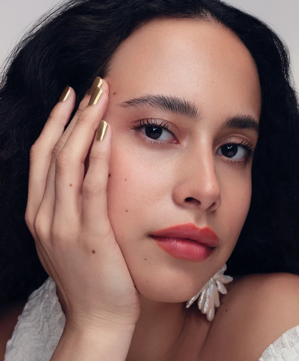 Woman with dark hair and gold nail polish touching her face against a neutral background
